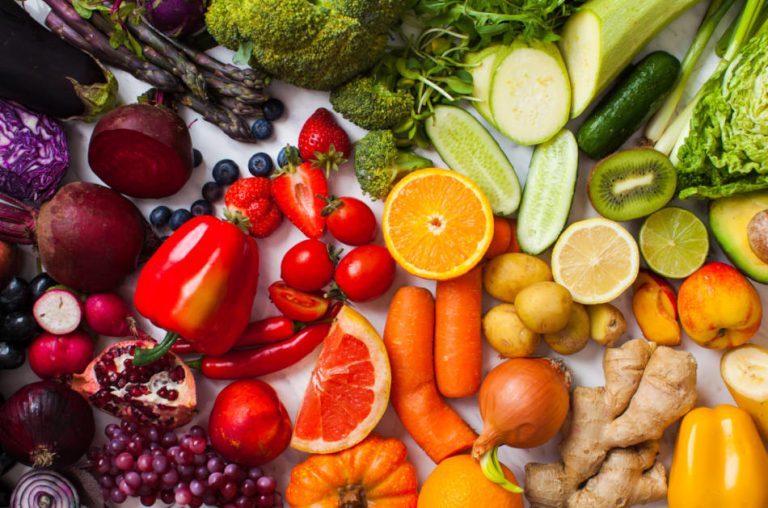 Rainbow-colored fruits and vegetables including purple cabbage and eggplant, red peppers and strawberries, orange carrots and citrus fruits, green onions and broccoli, and yellow peppers and squash.