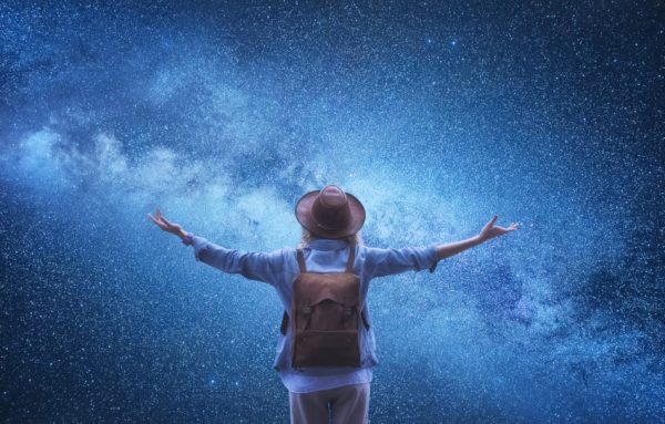 Female wearing hat and backpack with her back to the camera has arms outstretched in front of the night sky filled with stars.