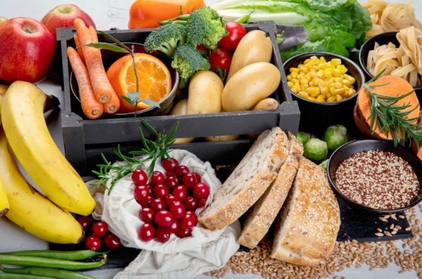 Best sources of carbs on a table, including bananas, apples, cherries, green beans, whole grain bread, potatoes, carrots, peppers, broccoli, tomatoes, corn, brussels sprouts, and sweet potatoes.