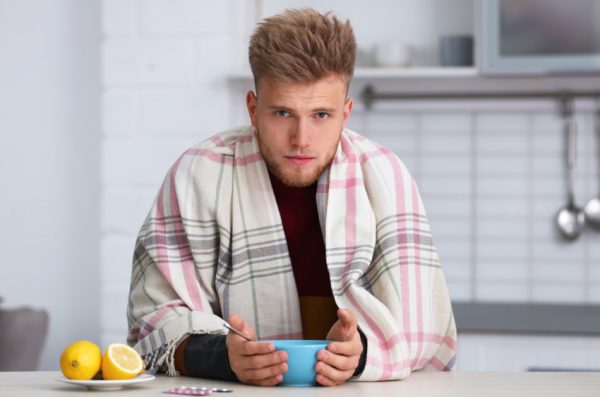 Sick young man eating soup to cure flu sits at the kitchen table with his hands on the bowl and a blanket draped over his shoulders.