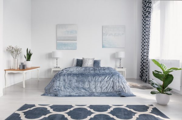 Elegant bedroom interior with big comfortable bed with blue bedding, paintings on the wall, and patterned carpet on the floor.