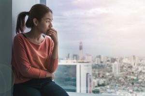 Depressed young Asian woman looking staring blankly out a window with the cityscape in the background.