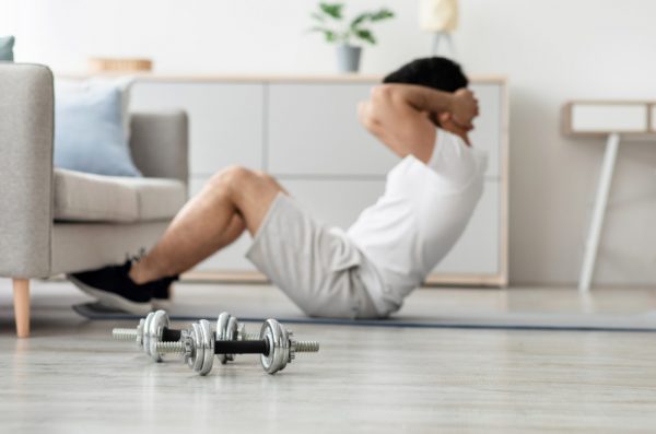 Millennial man doing abs exercises in morning in living room, with dumbbells sitting on floor in front of him.