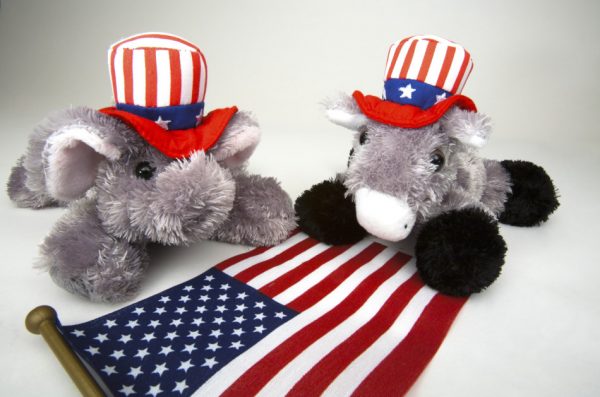 Elephant and donkey stuffed animals wearing red-white-and-blue hats sit on a table next to an American flag.
