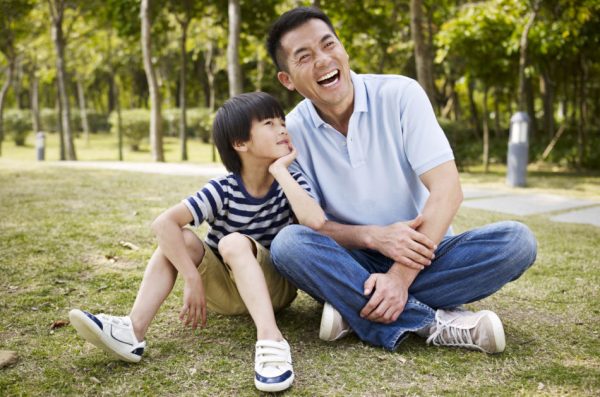 Asian father and young son sitting on the grass and laughing together.