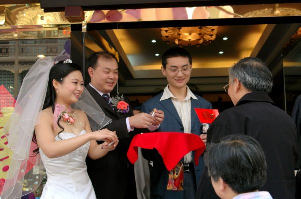 A bride and groom welcome guests to their wedding party luncheon by offering them cigarettes as they arrive at the restaurant on Qintai Road in Chengdu, China as one guest proffers a red hong bao (money) envelope gift.