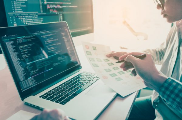 Two people sitting at a desk in an office with a laptop computer and a desktop computer, working together on a coding project.