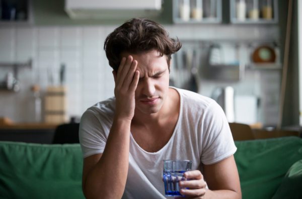 Young man sitting on a green sofa with a glass of water and his hand to his head, grimacing.