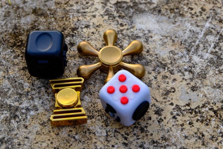 Four fidget toys including a black fidget cube, a white fidget cube with red and white spots, and two golden fidget spinners sit on a marble countertop.