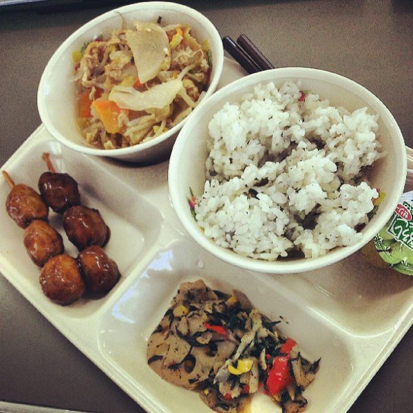Lunch tray from a school in Japan with a bowl of rice, a bowl of stir fried vegetables, some tofu mixed with red peppers, some barbeque meatballs on a stick, a juice cup, and chopsticks.
