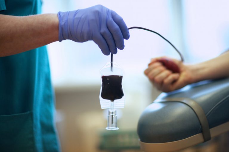 A gloved hand holds a bag filling with blood with the arm of the donor seen in the background.