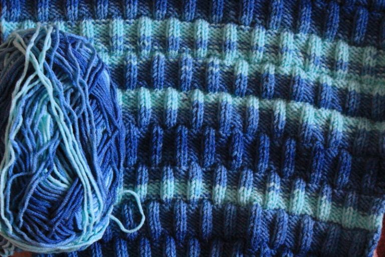 Closeup of knit and purl stitches in an alternating pattern with a skein of yarn in shades of blue ranging from very light to very dark.