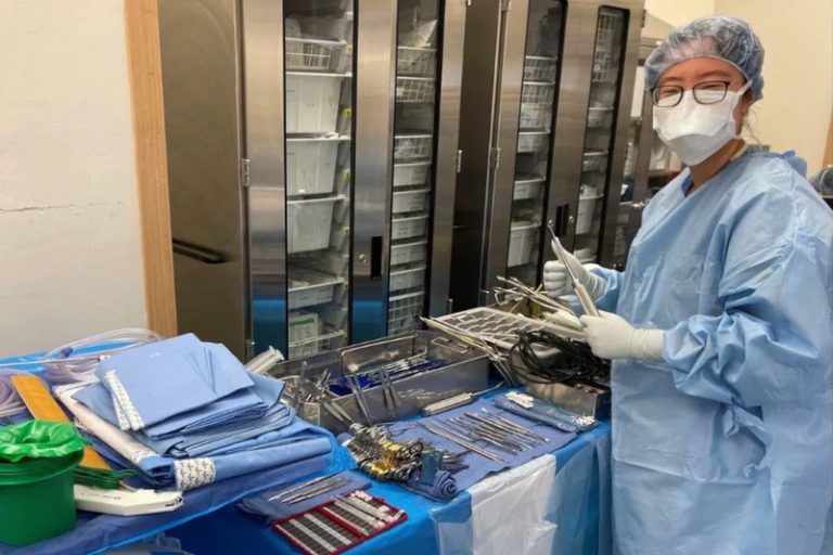 OR nurse in gloves, gown, and cap ready to assist in surgery stands by a tray laid out with instruments.