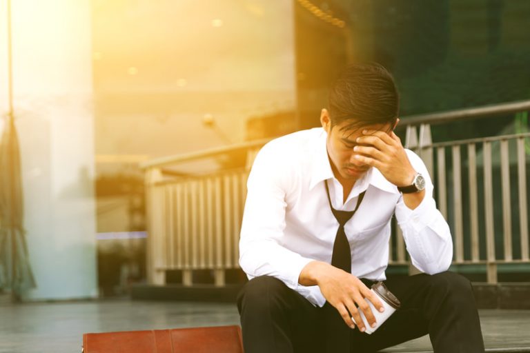 Worried Asian businessman sitting outside holding a dispossable cup while rubbing his temples.