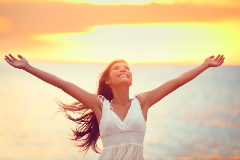 Joyful woman with head back and arms outstretched smiles while standing on the beach at dawn.