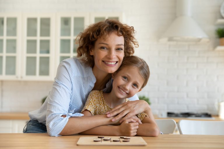 Mother with her arms around her daughter as she sits at the kitchen counter with a solved wooden puzzle in front of her.