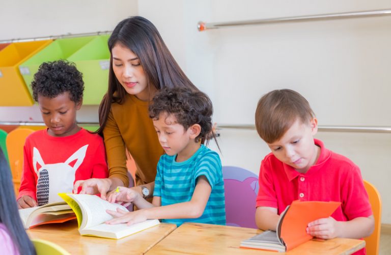 Female Asian teacher helping children with different ethnic backgrounds read a book.