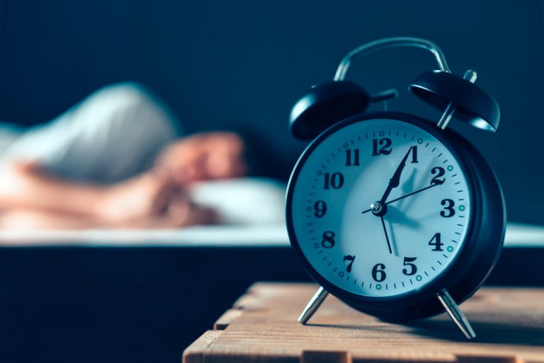 Alarm clock sitting on a bedside table shows the time at five minutes past five while a man is seen sleeping in bed in the blurred background.