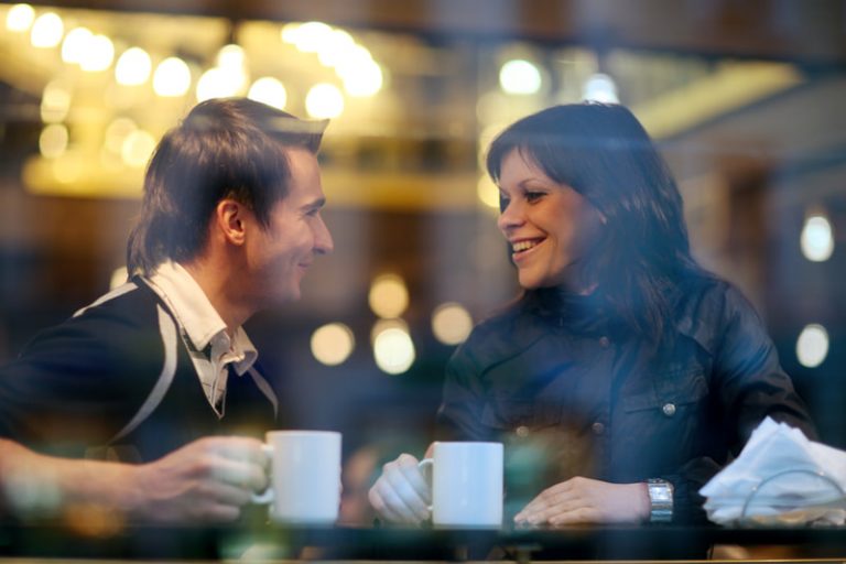 Happy young couple in a cafe smiling and talking, each with a beverage in a white mug.