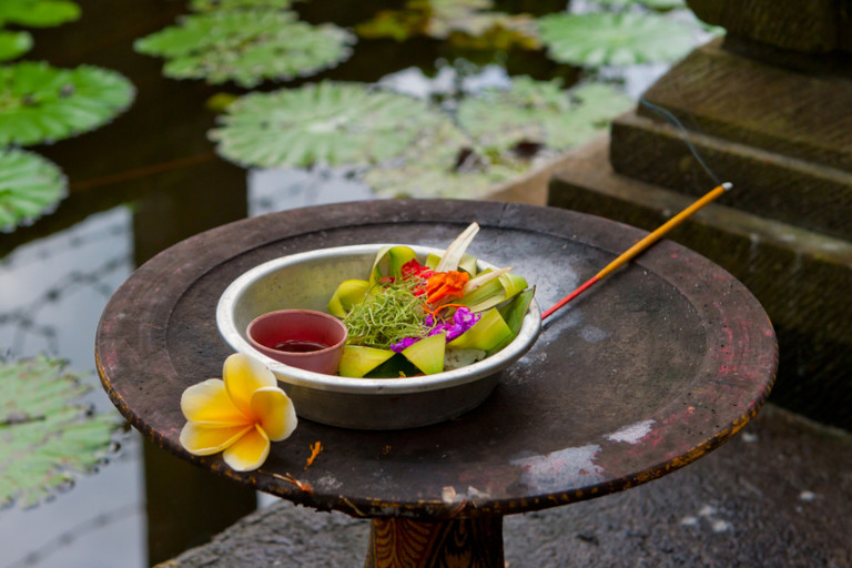 A bowl of colorful vegetables and a cup of wine sit next to a beautiful yellow flower and an incense stick given as a traditional Balinese offering at a shrine.