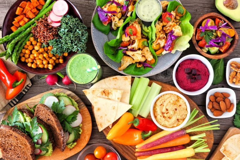 Healthy lunch table scene with nutritious bowl of garbanzo beans with kale, asparagus, sweet potatoes, and radishes beside a plate of lettuce wraps, some vegetables with humus, sandwiches, salad, and a green smoothie.