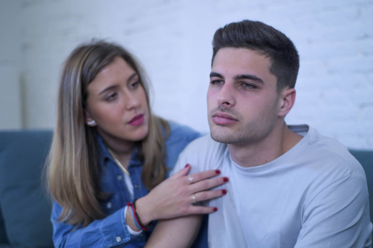 Sad young man sits on the couch as his wife tries to cheer him up.