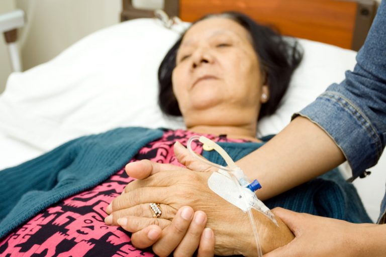 Elderly Asian female lying in bed with an IV inserted as a young woman holds her hand.