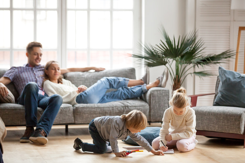 Husband sits on sofa while his wife stretches out and leans against him, watching their son and daughter who are playing together on the living room floor.