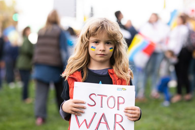 Blonde girl with Ukraine flags painted on both her cheeks holds a sign that says 'STOP WAR.'