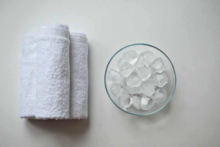 Ice cubes in a bowl sit next to 3 white cotton towels rolled up and stacked on top of each other.