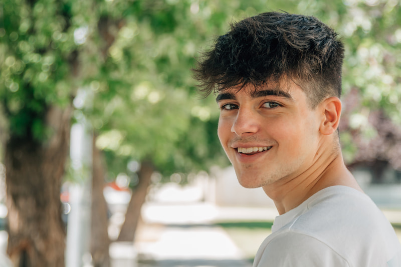 Smiling teen boy with a trendy haircut is standing outside on the sidewalk under a tree.