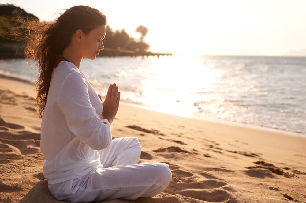 Woman wearing white sits on a beach with eyes closed, meditating.