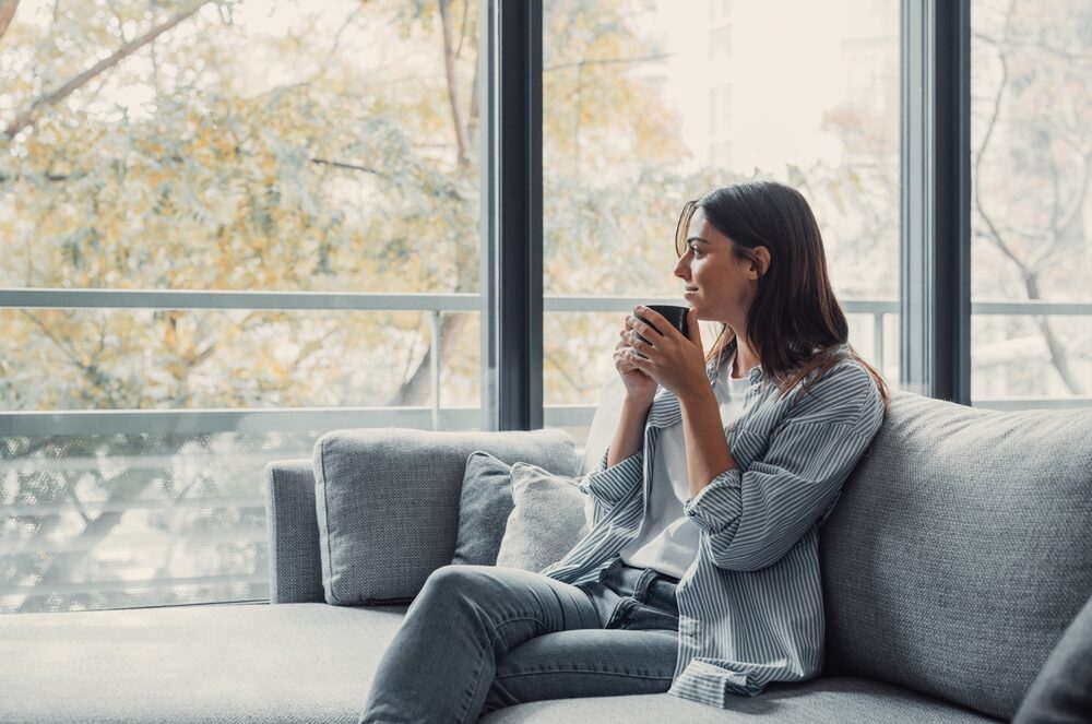 Young woman lost in thought sitting on a sofa looking out the window holding a mug with a warm beverage.