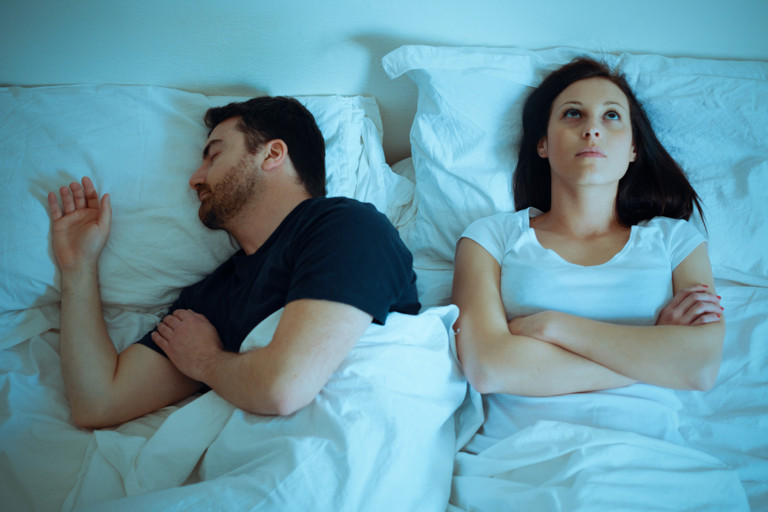 Man sleeping while upset woman lays in bed awake next to him.