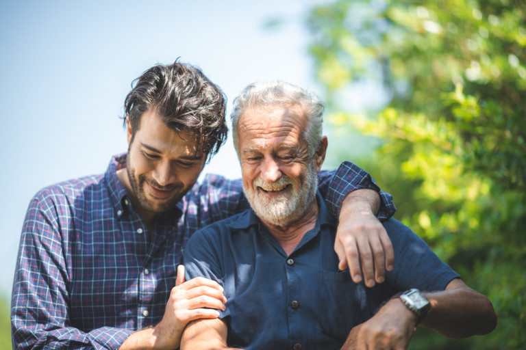 Adult son walking together with his arm around his elderly father outside as they both smile, enjoying a beautiful day.
