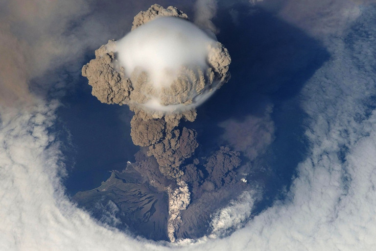Aerial view of a volcanic eruption with a massive ash plume billowing into the atmosphere, surrounded by clouds.