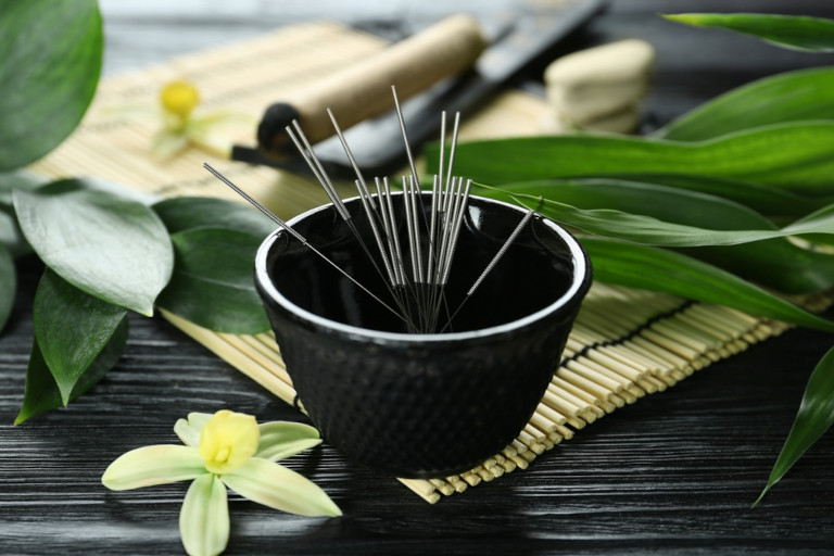 A bowl containing acupuncture needles sits on a bamboo mat.