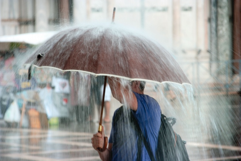 Man standing on a city sidewalk under a brown umbrella while it is raining heavily.