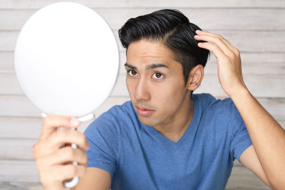 Asian male examining his hair in a hand-held mirror.
