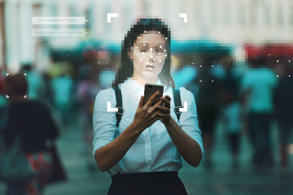 A woman stands in a crowded city street holding a smartphone while a facial recognition overlay maps her face, symbolizing digital surveillance and identity tracking in modern society.