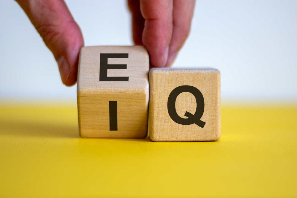 Two blocks with letters on them, one with vowels and the other with consonants, sitting on a yellow surface with a man flipping up the vowel block so it shows both 'E' and "I" in front of the letter 'Q'.