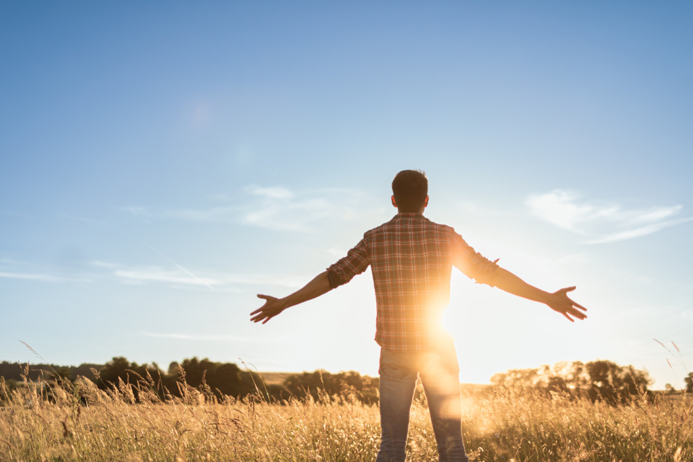 Young man standing in a field facing the sun with his arms out.