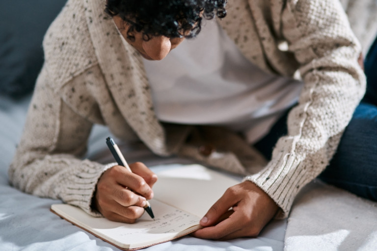 A young man on the floor with a pen and notebook, making a list.