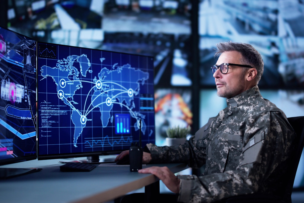 Image of a soldier in camouflage monitoring a world map and data dashboards on large screens in a military command center.