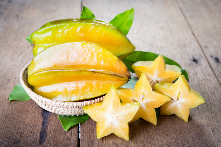 Whole starfruit in a basket sitting on green leaves next to cut starfruit all on a wooden surface.