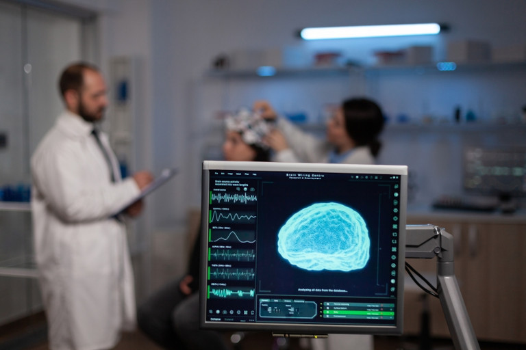 A screen showing brain activity being monitored in a neurology lab with a patient, technician, and physician in the background. 