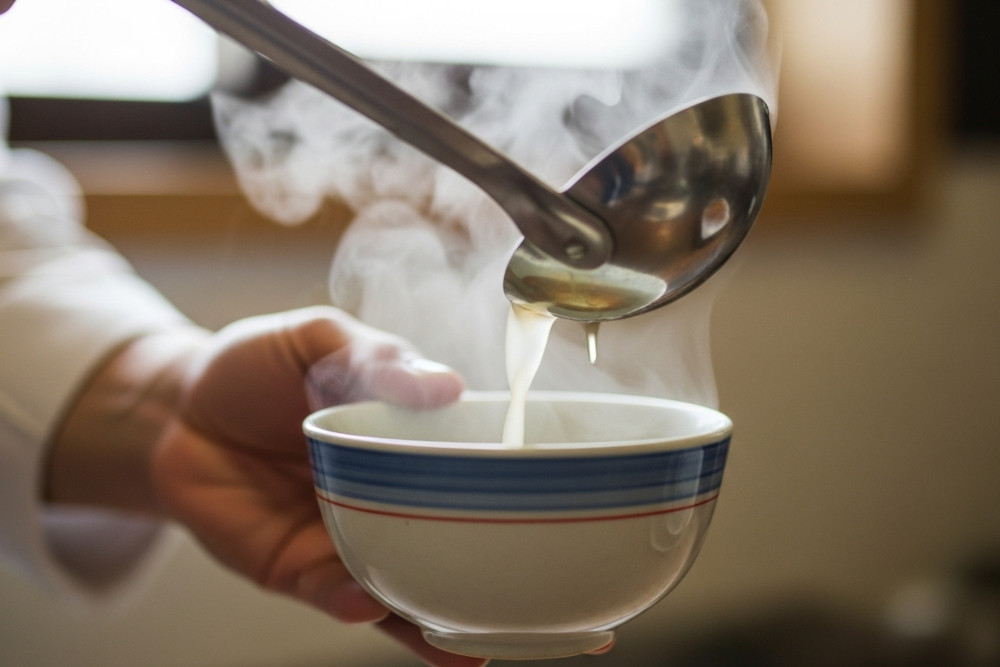 AI-generated image of hands ladling steaming soup into a simple bowl.