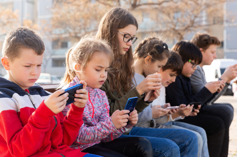A group of children sit side by side engrossed on their phones.