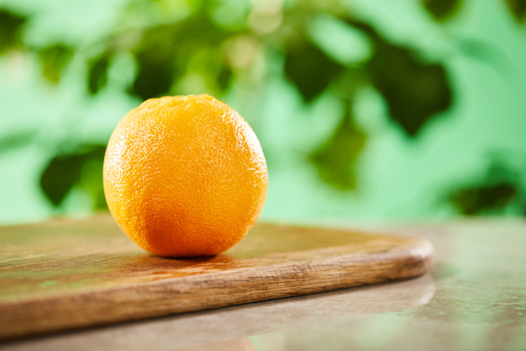 An orange sits on a cutting board while out-of-focus green leaves are seen in the background.