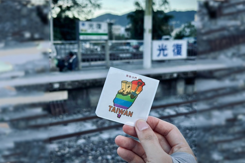 Volunteer holding a card showing Taiwan and a shovel sitting next to muddy boots.